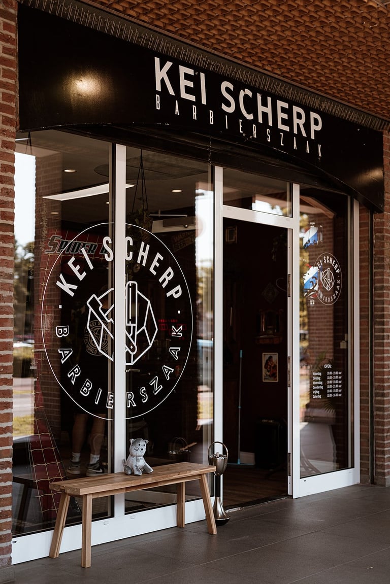 Modern brick barbershop storefront with Kei Scherp signage, white framed windows, and wooden bench seating outside