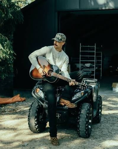 Man in baseball cap playing acoustic guitar while sitting on an ATV in a shadowy outdoor setting