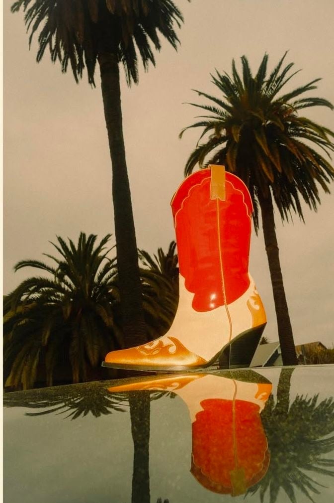 Red and gold western cowboy boot reflected in water with palm trees silhouetted in background