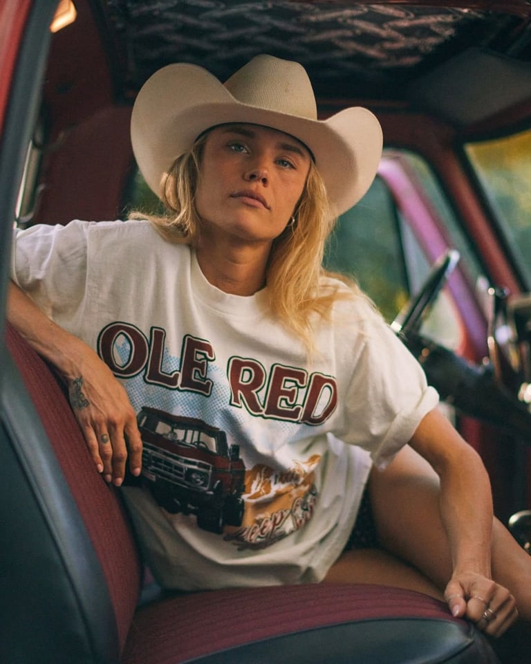 Woman in white graphic t-shirt and cream cowboy hat sitting in car, looking upward