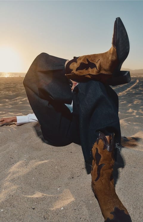 Person lying on beach sand wearing brown boots and dark clothing with legs up in playful pose at sunset
