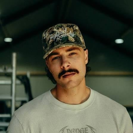 Man with mustache wearing camouflage baseball cap and light gray t-shirt, looking at camera in indoor setting