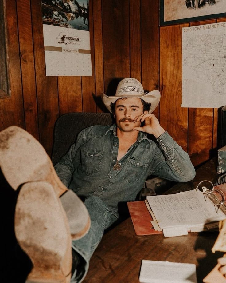 Man in cowboy hat and denim shirt sitting at wooden desk with papers and boots, in rustic wood-paneled room