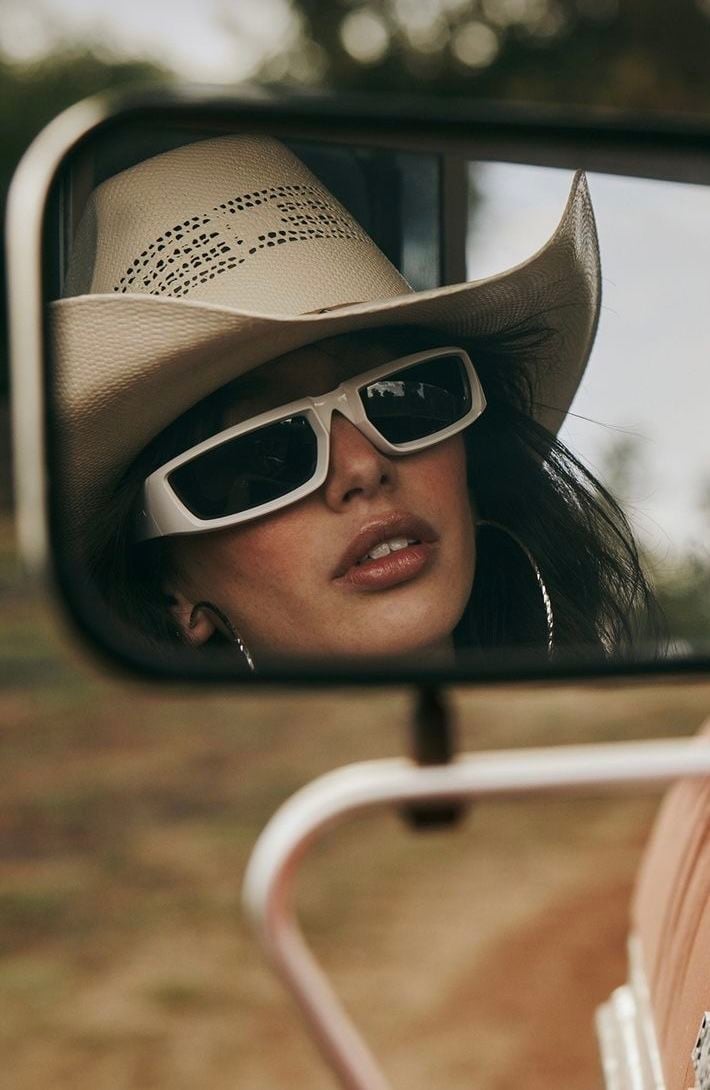 Woman in white cowboy hat and sunglasses reflected in car mirror against rural backdrop