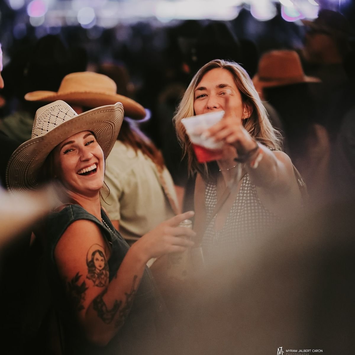 Two smiling women enjoying an outdoor event at night, one wearing a cowboy hat and showing a tattoo on her arm