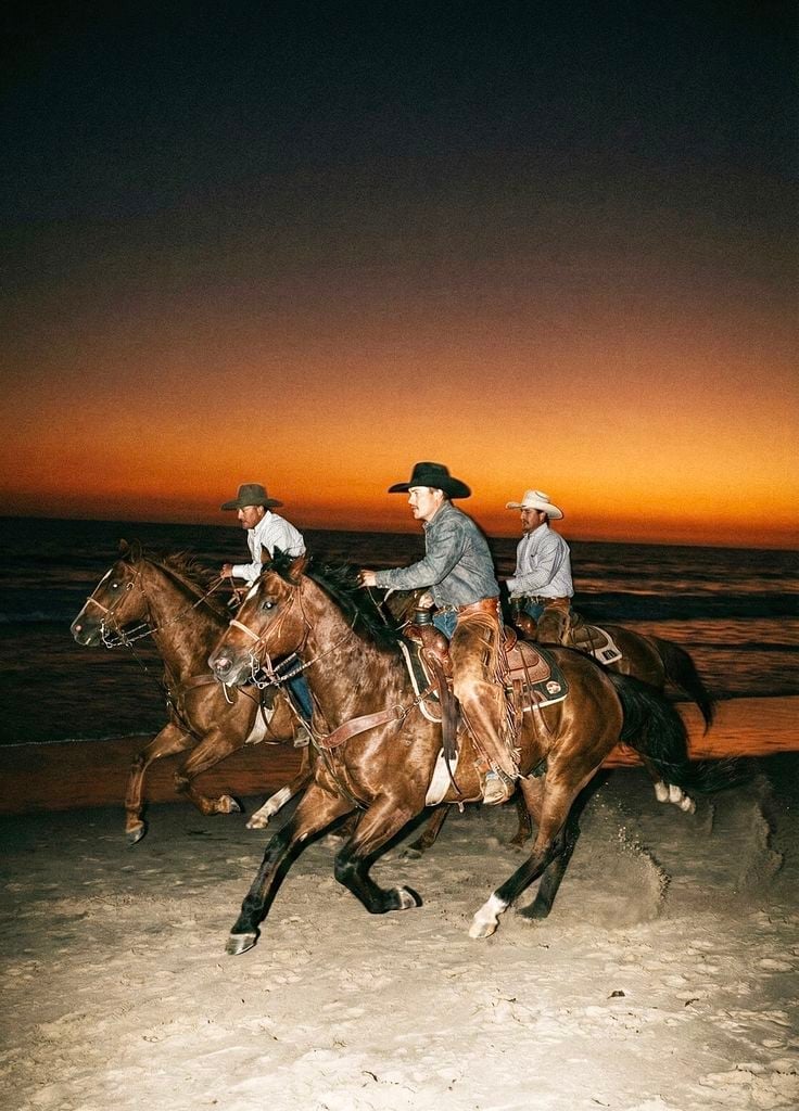 Three cowboys on horseback riding along a beach at sunset with an orange sky